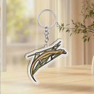 May include: A clear acrylic keychain featuring a printed illustration of a brown and tan lizard climbing on green grass blades. The lizard has a long tail and is attached to a silver-colored metal split ring by a short chain link connector.