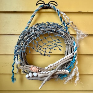 May include: A coastal-themed wreath with a driftwood base, adorned with a blue fishing net, rope, and seashells. A small crab figurine is placed on the driftwood. The wreath is suspended by a rope and a black metal hook.