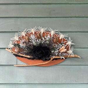 Puede incluir: Una decoración de Halloween con una base de madera circular en color naranja. Presenta arañas blancas, una rosa negra, follaje seco y una ramita. Diseñada para montaje en pared.