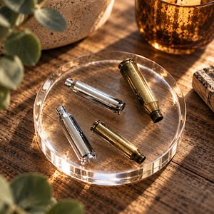 May include: Four cylindrical metal casings arranged on a clear circular acrylic tray. Two casings are finished in a polished silver tone, while the other two feature a weathered brass appearance. Each casing has a narrow neck and a hollow opening.