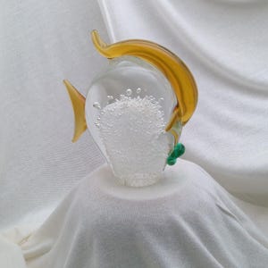 May include: A clear glass fish figurine featuring a central body filled with numerous small air bubbles. The fish has a curved amber-colored dorsal fin, a matching amber tail fin, and a small green glass accent near the bottom fin area.