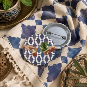 Op de afbeelding: Een ronde button met een speld aan de achterkant, voorzien van een blauw-wit geometrisch tegelpatroon. Het ontwerp toont twee handen die naar elkaar reiken, waarbij de ene een klein glas met amberkleurige vloeistof vasthoudt en de andere verse munt.