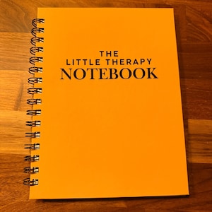 May include: A bright orange spiral-bound notebook with black wire binding along the left side. The front cover features the text THE LITTLE THERAPY NOTEBOOK printed in a bold, black, serif font centered on the page.