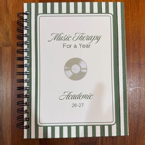 May include: A spiral-bound planner with a white and dark green striped border. The cover features the text "Music Therapy For a Year" and "Academic 26-27" in a serif font, with a centered illustration of a vinyl record in a muted olive green tone.