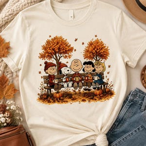 May include: A cream-colored short-sleeve t-shirt featuring a graphic print of the Peanuts characters sitting on a park bench under autumn trees. The characters are holding mugs, surrounded by fallen orange leaves and pumpkins on the ground.