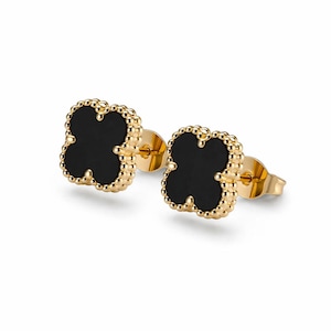 May include: A pair of stud earrings featuring a four-leaf clover shape with a smooth black center inlay. The clover motif is framed by a gold-toned beaded border, and each earring includes a matching gold-toned post and butterfly back closure.