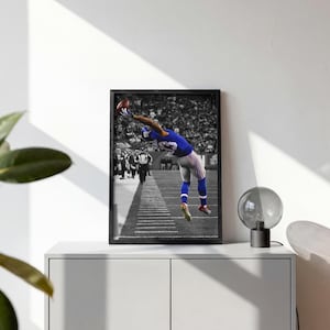 May include: A framed wall art print featuring a black and white photograph of a football player in a blue jersey leaping to catch a brown football. The player is mid-air on a field with yard lines visible, while the stadium crowd is blurred.