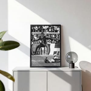 May include: A black and white framed wall art print depicting a football player in mid-air leaping over a downed opponent on a field. The action shot captures the dynamic movement of the athlete holding a football while the other player lies on the ground.