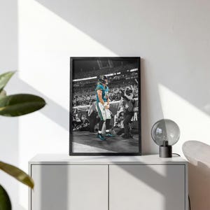 May include: A framed black and white photograph featuring a football player in a teal jersey standing on a field. The player wears a helmet and white pants. In the background, a photographer with a camera captures the scene amidst a stadium crowd.