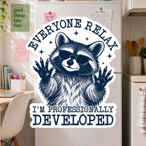 May include: A die-cut vinyl sticker featuring a dark blue illustration of a raccoon wearing sunglasses with its paws raised. The text reads EVERYONE RELAX I'M PROFESSIONALLY DEVELOPED in a bold, dark blue font against a white background.