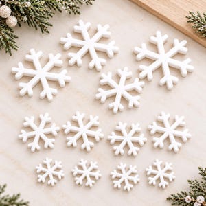 May include: A collection of twelve white snowflake ornaments arranged in three rows of varying sizes. Each snowflake features a six-pointed star design with branching arms, crafted from a smooth, solid material with a matte finish.