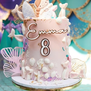 Puede incluir: Un pastel cilíndrico con glaseado rosa claro, decorado con conchas, estrellas de mar y colas de sirena de fondant blanco. El nombre Emma y el número 8 aparecen en letras doradas al frente. Una figura de sirena corona la parte superior.