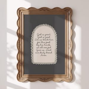 Puede incluir: Un cuadro rectangular en un marco de madera ondulado con tallas florales. El fondo gris oscuro presenta un arco central de color crema con un borde festoneado que contiene el texto: God is great. God is good. Let us thank him for this food. By his hands, we all are fed. Give us, Lord, our daily bread. Amen.