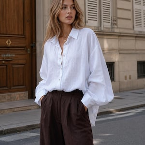 May include: A crisp white long-sleeved button-down shirt with a relaxed fit and a classic collar, paired with dark brown high-waisted trousers featuring a pleated front design and side pockets, creating a clean and structured two-piece outfit.