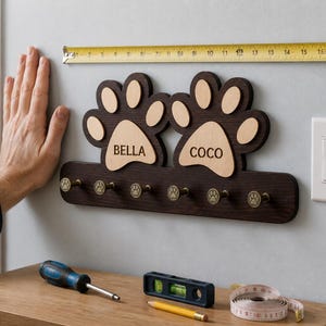 Puede incluir: Un perchero de pared de madera marrón oscuro con dos formas grandes de huellas de pata, con los nombres BELLA y COCO grabados en madera clara. El soporte incluye seis ganchos pequeños color latón, cada uno con una huella grabada.