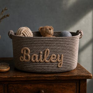 Pode incluir: Uma cesta redonda de corda trançada na cor taupe com o nome Bailey em letras de corda em relevo de tom mais claro na frente. A cesta possui duas alças de corda integradas nas laterais e contém um pequeno urso de pelúcia e uma bola.