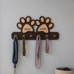 Puede incluir: Un soporte de pared para correas de mascotas hecho de madera marrón oscura, con dos recortes grandes en forma de huella en la parte superior grabados con los nombres MILO y LUNA. Incluye cinco ganchos de metal color latón antiguo.