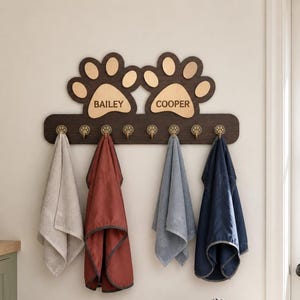 Puede incluir: Un perchero de pared de madera marrón oscuro con dos grandes formas de huellas de pata en la parte superior. La pata izquierda está grabada con el nombre BAILEY y la derecha con COOPER. Incluye siete ganchos pequeños color latón con forma de pata.
