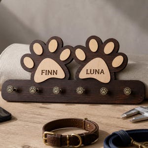 Puede incluir: Un perchero de pared de madera marrón oscuro con dos grandes formas de huellas de pata en la parte superior. Las almohadillas son de madera clara y están grabadas con los nombres FINN y LUNA. Seis ganchos metálicos con detalles de huellas.