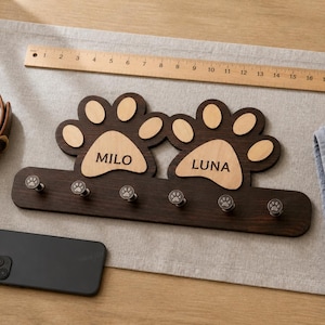 Puede incluir: Un perchero de pared de madera marrón oscuro con dos formas de huellas grandes con centros de madera clara grabados con los nombres MILO y LUNA. Tiene seis ganchos metálicos pequeños con detalles de huellas y mide 45 cm de ancho.
