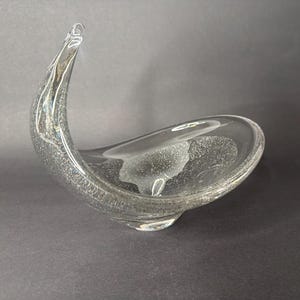 May include: A clear glass bowl with an asymmetrical, curved silhouette that rises to a high, pointed tip on one side. The glass features a dense, uniform distribution of tiny air bubbles throughout its structure, creating a textured appearance.