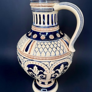 Può includere: Una brocca in ceramica con base color crema e accenti blu scuro e marrone. Il recipiente presenta intricati motivi in rilievo, tra cui disegni floreali, reticoli geometrici e scanalature verticali, con un singolo manico curvo.