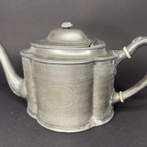 Op de afbeelding: Een metallic grijze tinnen theepot met een geschulpt lichaam, een gebogen tuit en een deksel met een kleine knop. Het handvat is bevestigd met twee lichtgekleurde ringen en heeft een sierlijk, gebogen ontwerp. Het oppervlak heeft horizontale lijnen.