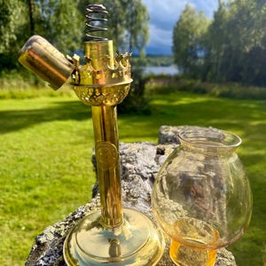 Op de afbeelding: Een koperen olielampvoet met een ronde standaard, een verticale steel en een kroonvormige brander met een zichtbare metalen veer. Naast de voet staat een losse, bolvormige, amberkleurige glazen cilinder met een wijd uitlopende opening.