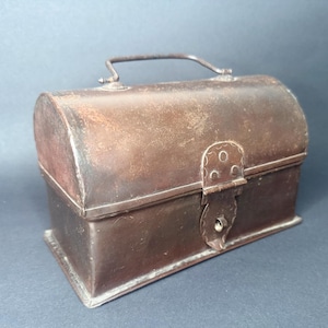 Op de afbeelding: Een rechthoekige metalen doos met een afgerond, gewelfd deksel en een verweerde, donkerbruine patina. De container is voorzien van een scharnierend deksel, een metalen handvat en een decoratief sluitmechanisme met een sleutelgat aan de voorzijde.