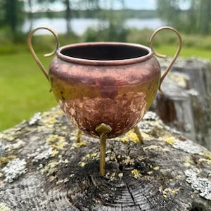 Pode incluir: Um pequeno pote de cobre com corpo arredondado, apresentando duas alças de metal finas e curvas nos lados opostos. O pote é sustentado por três pernas de latão finas e pontiagudas fixadas na base, exibindo um acabamento metálico desgastado.