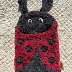 Könnte beinhalten: Ein Marienkäfer-Waschhandschuh aus rotem und schwarzem Frotteestoff. Der Handschuh hat einen schwarzen Kopf mit zwei Antennen, gestickten weißen Augen und einen roten Körper mit schwarzen Punkten. Ein kleines Stoffetikett ist seitlich angebracht.