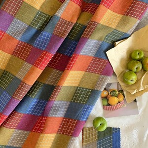 May include: A fabric featuring a multicolored plaid pattern with red, orange, yellow, green, blue, and purple squares. The textile has a white grid overlay and small white dots. Nearby are green plums on paper and a card with the text Bonheur Joie.