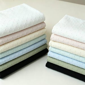 May include: Two stacks of folded fabric featuring a Swiss dot texture. The colours in each stack include white, light pink, cream, light blue, sage green, and black. The fabric has a consistent raised dot pattern across the entire surface.