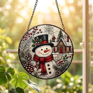 May include: A circular stained glass suncatcher featuring a snowman wearing a black top hat and a red scarf. The scene includes a winter cabin, pine cones, red berries, and snowflakes, all framed in a dark metal border with a hanging chain.