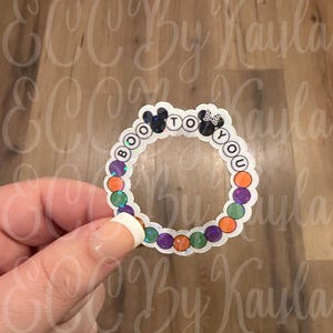 May include: A circular sticker featuring the text BOO TO YOU in black letters. The design includes two mouse-ear silhouettes and a border of alternating orange, purple, and green holographic circles. The sticker has a white scalloped edge.