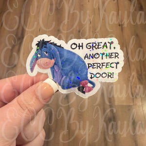 May include: A die-cut sticker featuring a blue donkey character with a pink bow on its tail, accompanied by the black text "OH GREAT, ANOTHER PERFECT DOOR!" The sticker has a holographic finish that reflects light with small, colourful sparkles.