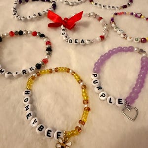 May include: A collection of handmade beaded bracelets featuring various color schemes, letter beads spelling out words like HONEYBEE, PURPLE, and VAMPIRE, and decorative charms including a bee, a heart, and a red ribbon bow on a white surface.