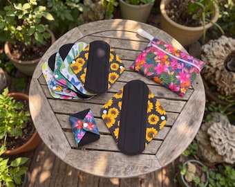 May include: A collection of reusable cloth menstrual pads featuring various patterns including sunflowers, cacti, and tropical flowers, alongside a matching zippered storage pouch. The pads have black fabric centers and white snap closures.