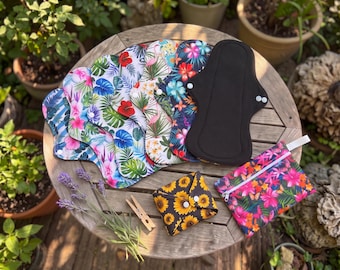 May include: A collection of reusable cloth menstrual pads featuring various floral and tropical prints, one solid black pad, a sunflower-patterned folded pad, and a matching zippered pouch, all arranged on a wooden surface.