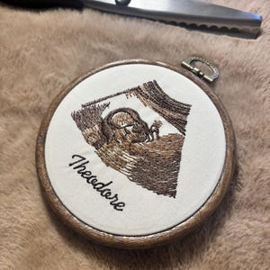 Könnte beinhalten: Ein runder Stickrahmen aus Holz mit cremefarbenem Stoff, auf dem ein Ultraschallbild eines Fötus in braunem Garn gestickt ist, sowie der Name Theodore in schwarzer Schreibschrift. Darüber liegt eine Metallschere mit gezackten Klingen.