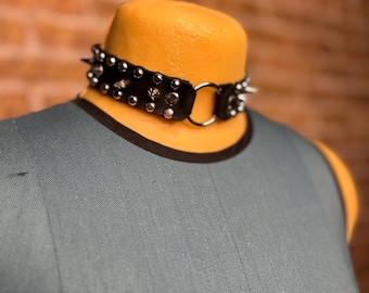 May include: A black leather choker necklace featuring two rows of silver-toned metal studs and a central silver-toned metal O-ring. The choker is displayed on a mannequin neck, showcasing the metallic hardware and the dark material construction.