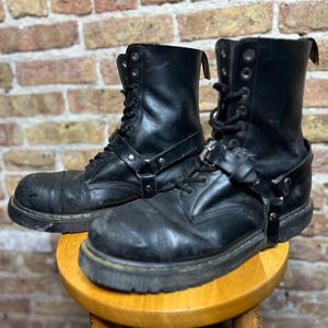 May include: A pair of black leather lace-up boots featuring a rounded toe, a thick rubber sole with yellow stitching, and a decorative harness strap with a silver-toned metal buckle wrapped around the ankle and mid-foot area of each boot.