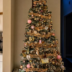 May include: A flocked Christmas tree topped with a brown pointed hat, decorated with warm white string lights, gold and pink ornaments, and tan ribbons printed with the text: I solemnly swear that I am up to no good and Mischief managed.