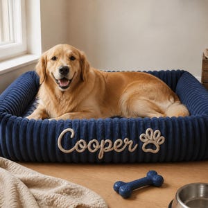 May include: A rectangular navy blue corduroy dog bed with raised sides. The front of the bed features the name Cooper embroidered in white script lettering next to a white embroidered paw print. A golden retriever is lying inside the bed.