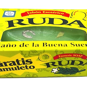 May include: A rectangular yellow box containing a green bar of soap with a small silver horseshoe charm embedded inside. The packaging features the text RUDA, Jabón Esotérico, Baño de la Buena Suerte, and Gratis amuleto, along with illustrations of horseshoes and a green sprig.