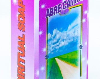 May include: A rectangular box of Abre Camino spiritual soap with a bright purple and white design. The front features an illustration of a dirt road leading through a green field under a blue sky with clouds. Text includes JABON, SPIRITUAL SOAP, ABRE CAMINO, OPEN ROAD, and For External Use Only.