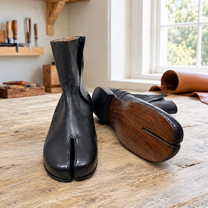 May include: A pair of black leather tabi boots featuring a distinct split-toe design. One boot stands upright, while the other lies on its side, revealing a dark brown wooden sole with a visible split that matches the toe separation of the boot.