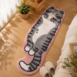 Puede incluir: Una alfombra larga y rectangular con forma de gato acostado boca arriba. La alfombra presenta un patrón de gato atigrado gris y blanco con una superficie suave y texturizada. El gato tiene una barriga blanca, extremidades a rayas grises y una cola larga, todo bordeado por un borde rosa.