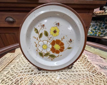 Puede incluir: Un plato redondo de cerámica con una superficie de color blanco roto moteado y un borde marrón sólido. El centro presenta una ilustración floral con flores amarillas, naranjas y blancas, hojas verdes y tallos marrones finos sobre un fondo claro.