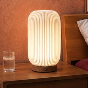 May include: A cylindrical table lamp with a white, vertically ribbed shade that emits a soft, warm glow. The lamp features a circular wooden base with a small black power button on the side, providing a steady light source for a bedside table.